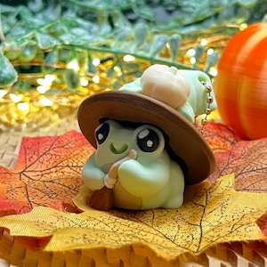 May include: A whimsical green frog figurine wearing a brown witch hat and holding a tiny broom. The frog has large, expressive eyes and is set on a bed of autumn leaves. A small pumpkin is also visible.
