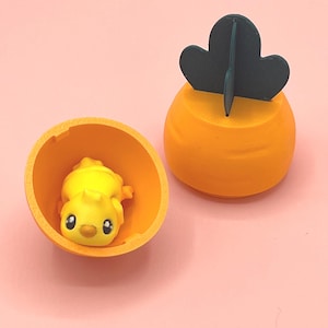 Può includere: Un contenitore a forma di carota arancione con un dettaglio di foglia verde scuro, accanto a un guscio d'uovo arancione abbinato contenente una figura di pulcino giallo con grandi occhi. Su sfondo rosa chiaro.