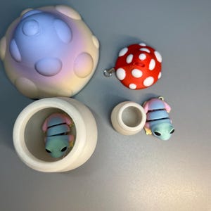 Może przedstawiać: Zestaw trzech miniaturowych figurek w kształcie grzyba. Jedna to duży, biały grzyb z fioletowym i r&oacute;żowym gradientem. Druga to mniejszy, czerwony grzyb w białe kropki. Trzecia to mała, niebieska i r&oacute;żowa figurka żaby.