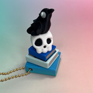 Pode incluir: Um colar com pingente com um corvo preto empoleirado em cima de uma caveira branca, que repousa sobre uma pilha de livros azuis e azul claro. O colar tem uma corrente de bolas dourada.