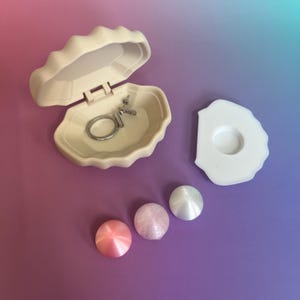 Peut inclure: Une boîte à bijoux en forme de coquillage de couleur crème est ouverte, révélant une bague argentée. Trois sphères nacrées roses et blanches sont à côté d'un couvercle blanc en forme de coquillage. Le fond est un dégradé de violet et de bleu.
