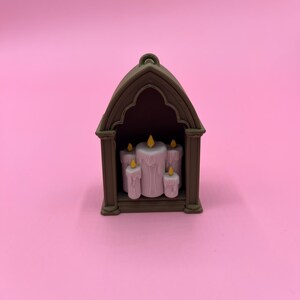 以下が含まれることがあります： 茶色の小さなアーチ型の構造の中に、黄色い炎のついた5本のピンク色のろうそくが収められています。ろうそくのサイズは様々で、中央のろうそくが一番大きいです。背景は単色のピンク色です。