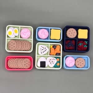 May include: A collection of colorful, miniature bento boxes. Each box is divided into compartments, filled with various food items like cookies, eggs, cheese, and fruit. The boxes are in shades of pink, blue, green, and navy.