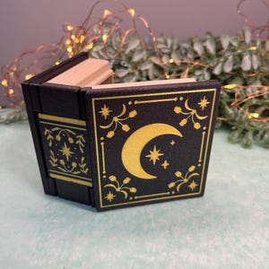 May include: A small, black box with a hinged lid, resembling a book. The front cover features a gold crescent moon and stars design. The box is decorated with gold floral and star accents. The interior is made of light-coloured wood.