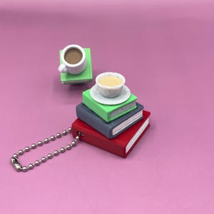 Pode incluir: Um chaveiro com uma pilha de livros em miniatura vermelhos, cinzentos e verdes, encimada por uma pequena chávena de café. Uma segunda chávena de café em miniatura está em cima de um livro verde. O chaveiro tem uma corrente de bolas prateada.