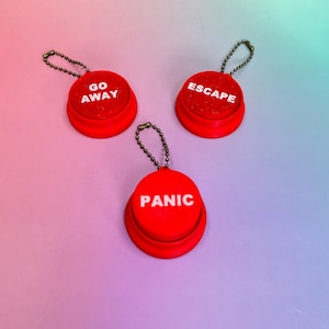 Könnte beinhalten: Drei rote Neuheitsknöpfe mit silbernen Kettenanhängern. Jeder Knopf hat ein anderes weißes Wort: "GO AWAY", "ESCAPE" und "PANIC". Die Knöpfe befinden sich auf einem Farbverlaufshintergrund aus Rosa, Lila und Blau.