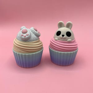 Puede incluir: Dos figuras decorativas con forma de cupcake. Una presenta un gato gris con huellas de patas rosas, la otra un conejito blanco. Ambos tienen glaseado rosa y beige, y moldes azules y morados. Sobre un fondo rosa.