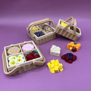 Op de afbeelding: Miniatuur picknickmanden gevuld met etenswaren, waaronder koekjes, aardbeien en kaas. De manden zijn beige met witte randen en hebben een handvat. Andere etenswaren liggen verspreid rond de manden op een paars oppervlak.