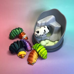 Peut inclure: Un ensemble de six figurines articulées de cloportes de différentes couleurs, dont vert métallique, orange, noir, violet et une blanche à taches noires, reposant dans une structure de grotte en plastique gris avec une base vert clair.