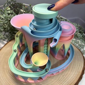 May include: A pastel rainbow colored marble run toy with a tower and a funnel. The toy is made of plastic and has a smooth, glossy finish.