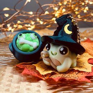 May include: A pair of whimsical Halloween-themed figurines. One is a light green frog in a cauldron with a bone, and the other is a light green creature wearing a black witch hat with a yellow crescent moon. The figurines are on a bed of autumn leaves.