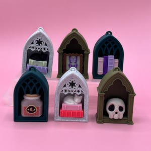 以下が含まれることがあります： さまざまな色のミニチュアゴシック様式の装飾品のコレクション。各装飾品は、本、猫、スカル、額入りの蝶など、さまざまなインテリアデザインが特徴です。装飾品はピンク色の背景に配置されています。
