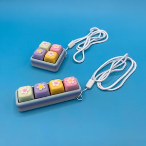 Puede incluir: Dos llaveros de colores pastel con un diseño de bandeja cuadrada, cada uno con cuatro bloques cuadrados con diseños de flores. Los bloques son en tonos de verde, morado, rosa y amarillo. Cada llavero tiene un cordón blanco.