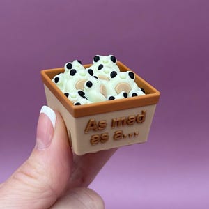 May include: A small, tan square container filled with cream-colored, cartoon-like creatures with black spots. The container has a brown rim and the text "As mad as..." in brown.
