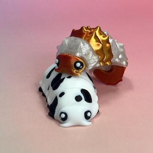 Peut inclure: Un jouet chenille imprimé en 3D avec un corps blanc et des taches noires. Il a une petite tête orange et une structure plus grande, multicolore et épineuse sur le dos. Le jouet est posé sur un fond dégradé rose et pêche.