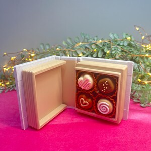 Op de afbeelding: Een klein, open boek met een doosje chocolaatjes erin. De chocolaatjes zijn hartvormig en rond, met witte, bruine en roze glazuur. Het boek heeft een witte kaft en beige pagina's. De achtergrond is roze en groen.