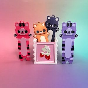 May include: A collection of colorful cat-shaped bookmarks and a decorative container. The bookmarks are in pink, orange, purple, and navy blue, each with a unique design. The container is white with a pink square featuring a strawberry design.