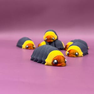 Op de afbeelding: Verschillende 3D-geprinte rupsfiguren. Elke rups heeft een grijs lichaam, een gele kop en oranje details. De figuren zijn gerangschikt op een roze oppervlak, waarbij sommige scherp en andere wazig zijn.