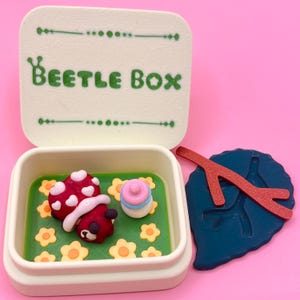 Puede incluir: Una caja rectangular blanca con la inscripción "BEETLE BOX" en verde. Dentro, una figura de mariquita roja y blanca descansa sobre una base verde con flores amarillas. También hay una hoja azul y una rama roja.
