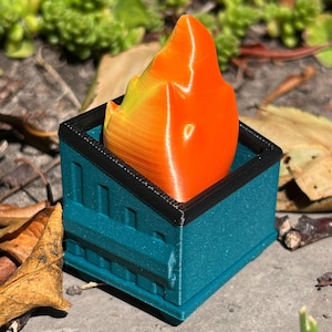 May include: A teal-colored, square container with a black rim, holding a flame-shaped object. The flame transitions from yellow to orange. The container has a textured surface and is sitting on a stone surface with leaves.
