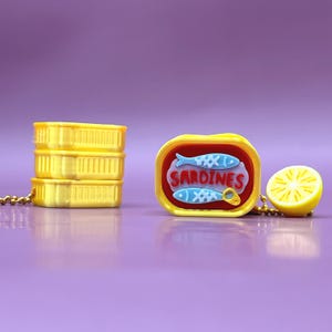 Op de afbeelding: Een grillige sleutelhanger set met drie gele sardineblikjes en een sardineblikje bedel met het woord "Sardines" en vis illustraties. Een gele citroen schijf bedel is ook inbegrepen, alles aan een gouden ketting.