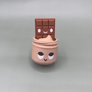 Può includere: Una statuetta stampata in 3D raffigurante un barattolo marrone chiaro con un volto sorridente e grandi occhi, contenente un ciuffo di crema beige. Una tavoletta di cioccolato marrone con un volto sorridente e occhi bianchi è posizionata nel barattolo.