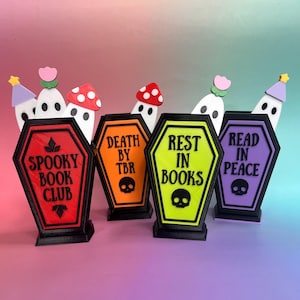 Peut inclure: Quatre porte-stylos en forme de cercueil rouge, orange, jaune fluo et violet, avec du texte noir et une icône de crâne. Le texte indique SPOOKY BOOK CLUB, DEATH BY TBR, REST IN BOOKS et READ IN PEACE. Chaque support contient un fantôme ou un champignon.