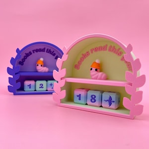 May include: Two decorative bookends in pink, purple, and green. Each bookend has a shelf with a worm figurine wearing a hat. The text "Books read this year" is arched above the shelf. Below are numbered blocks.