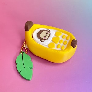 May include: A yellow banana-shaped silicone case featuring a white monkey face graphic and yellow keypad buttons. The case includes a gold-toned ball chain attached to a green leaf-shaped charm, providing a playful and functional accessory design.
