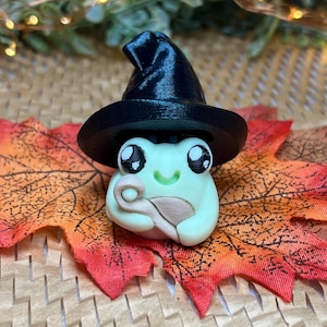 May include: A whimsical, handmade frog figurine wearing a black witch hat. The frog is light green with large, expressive eyes and a small smile. It rests on a colorful, autumn-themed leaf, creating a charming, seasonal display.