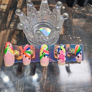 May include: A set of six artificial nails with a light pink base, each adorned with colorful abstract designs. The designs include stripes, dots, and floral patterns in shades of green, yellow, blue, and pink. A clear, crown-shaped holder sits above the nails.