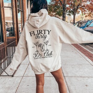 May include: A white hoodie with a black graphic that says "FLIRTY thirty EST. 1994 30's Club AGED TO PERFECTION". The graphic also includes a drawing of three cocktail glasses.