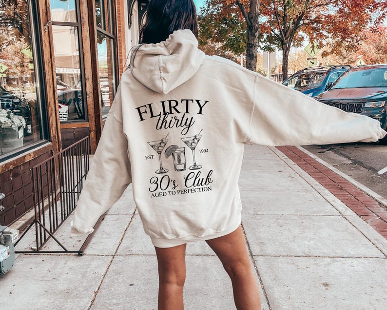 May include: A white hoodie with a black graphic that says "FLIRTY thirty EST. 1994 30's Club AGED TO PERFECTION". The graphic also includes a drawing of three cocktail glasses.