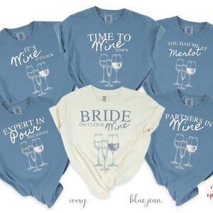 May include: Six blue jean and ivory t-shirts with wine-themed text and graphics. The shirts feature text such as "It's Wine O'Clock", "Time to Wine Down", "You Had Me at Merlot", "Expert in Pour Decisions", "Bride on Cloud Wine", and "Partners in Wine".
