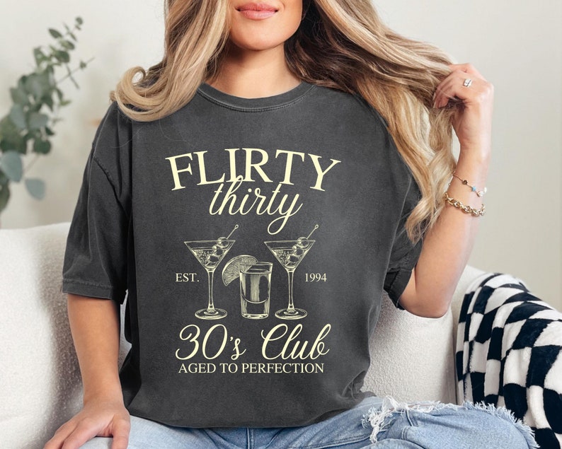 May include: A dark gray t-shirt with the text "FLIRTY thirty EST. 1994 30's Club AGED TO PERFECTION" and a graphic of three cocktail glasses.