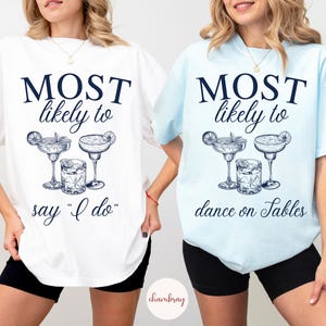 May include: Two white t-shirts with blue text and illustrations of cocktails. The text on the first shirt reads "MOST likely to say "I do"". The text on the second shirt reads "MOST likely to dance on tables".