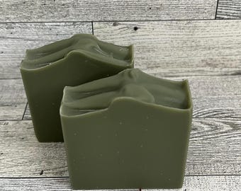 May include: Two olive green soap bars with a textured top are displayed. The soap bars are rectangular with a slightly uneven surface. The background is a light-colored wooden surface.