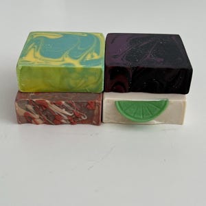 May include: Four handmade soap bars in a stack. The top bar is green and yellow, the next is purple and black, the third is red and white, and the bottom bar is white with a green lime slice.
