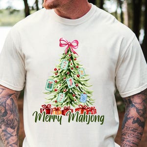 Pode incluir: Camiseta creme com um desenho de &aacute;rvore de Natal com la&ccedil;o rosa, enfeites e presentes. A &aacute;rvore &eacute; decorada com pe&ccedil;as de Mahjong e bengalas doces. O texto "Merry Mahjong" est&aacute; escrito em verde.