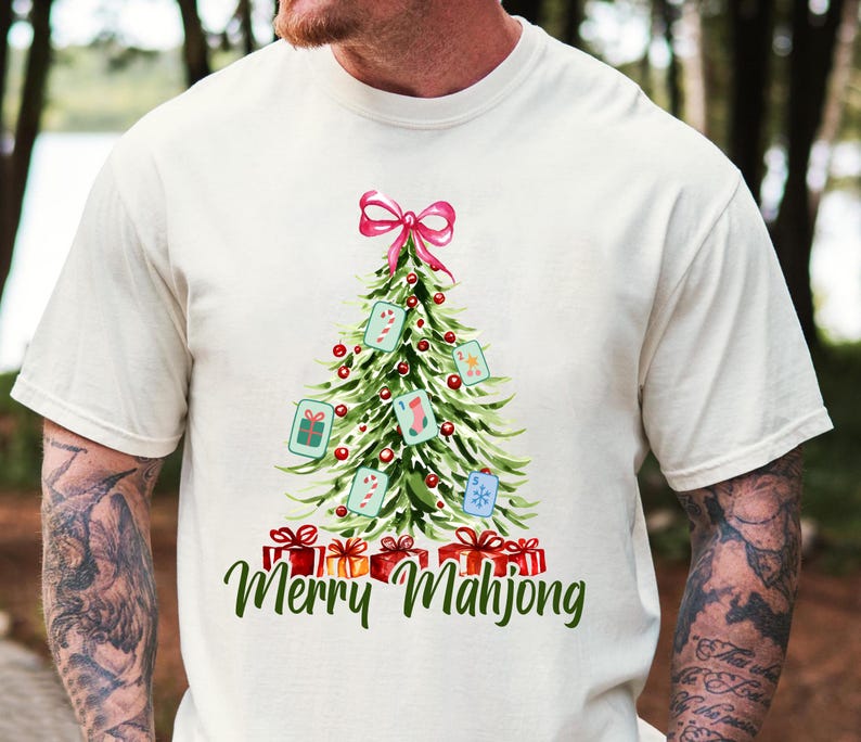 Pode incluir: Camiseta creme com um desenho de &aacute;rvore de Natal com la&ccedil;o rosa, enfeites e presentes. A &aacute;rvore &eacute; decorada com pe&ccedil;as de Mahjong e bengalas doces. O texto "Merry Mahjong" est&aacute; escrito em verde.