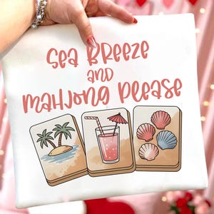 Może przedstawiać: Biała kwadratowa poduszka z r&oacute;żowym napisem "Sea Breeze and Mahjong Please". Poniżej trzy ilustrowane kafelki do mahjonga: tropikalna wyspa, r&oacute;żowy nap&oacute;j i muszle.