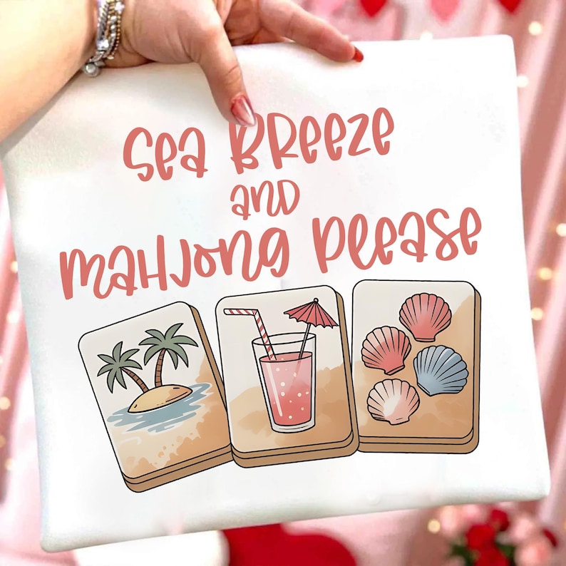 Może przedstawiać: Biała kwadratowa poduszka z r&oacute;żowym napisem "Sea Breeze and Mahjong Please". Poniżej trzy ilustrowane kafelki do mahjonga: tropikalna wyspa, r&oacute;żowy nap&oacute;j i muszle.