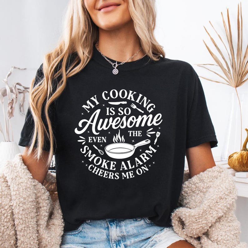 Puede incluir: Camiseta negra con un gr&aacute;fico blanco que dice: "My Cooking Is So Awesome Even The Smoke Alarm Cheers Me On." El dise&ntilde;o incluye una sart&eacute;n, llamas y utensilios. Una prenda informal.