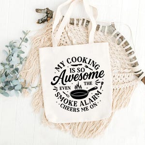 Puede incluir: Una bolsa de mano de lona blanca con el texto "MY COOKING IS SO Awesome EVEN THE SMOKE ALARM CHEERS ME ON." El dise&ntilde;o presenta una sart&eacute;n, utensilios y llamas en negro. La bolsa tiene asas beige.