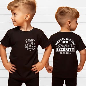 May include: Black t-shirts with white text and graphics. One shirt features a shield design with the name "Henry" and the words "Bride Security" around two rings. The other shirt has "Michael Wedding" and "Bride Security" with a date.
