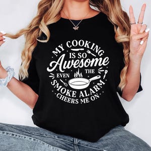 Puede incluir: Camiseta negra de cuello redondo con el texto blanco "MY COOKING IS SO Awesome EVEN THE SMOKE ALARM CHEERS ME ON" en un dise&ntilde;o circular. El dise&ntilde;o incluye una sart&eacute;n, utensilios y estrellas.