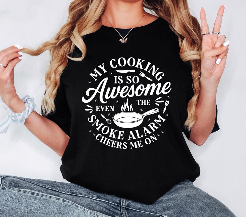 Puede incluir: Camiseta negra de cuello redondo con el texto blanco "MY COOKING IS SO Awesome EVEN THE SMOKE ALARM CHEERS ME ON" en un dise&ntilde;o circular. El dise&ntilde;o incluye una sart&eacute;n, utensilios y estrellas.