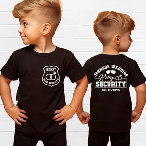 Op de afbeelding: Twee zwarte T-shirts met witte tekst. Het voorste shirt zegt "Henry Ring Security" met een badge en twee ringen. Het achterste shirt zegt "Johnson Wedding Ring Security 08-17-2025".