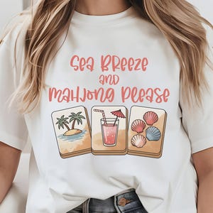 Może przedstawiać: Biała koszulka z r&oacute;żowym napisem "Sea Breeze and Mahjong Please". Koszulka przedstawia trzy ilustrowane kafelki: scenę plażową z palmami, r&oacute;żowy nap&oacute;j z parasolem i muszle. Projekt ma letni, wakacyjny motyw.