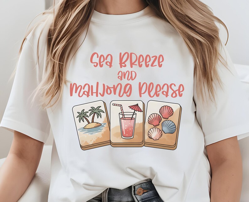 Może przedstawiać: Biała koszulka z r&oacute;żowym napisem "Sea Breeze and Mahjong Please". Koszulka przedstawia trzy ilustrowane kafelki: scenę plażową z palmami, r&oacute;żowy nap&oacute;j z parasolem i muszle. Projekt ma letni, wakacyjny motyw.